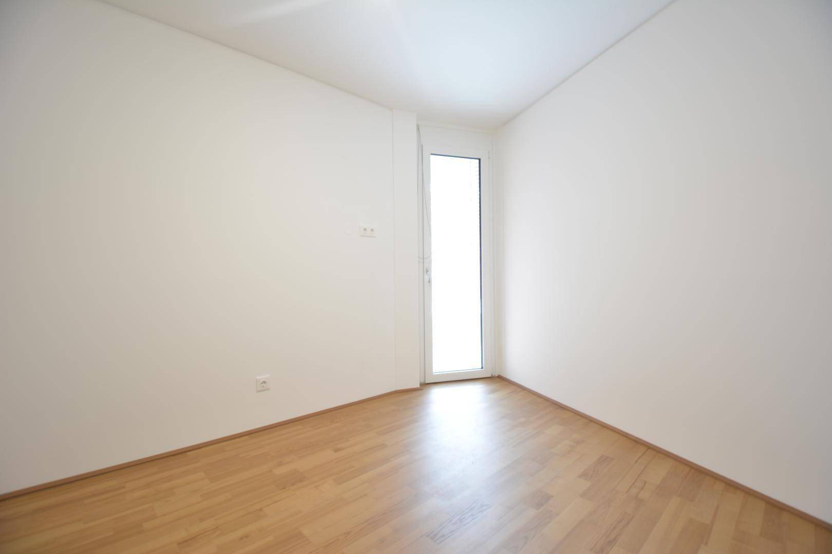 Helles Zimmer mit Parkettboden und einer hohen Fenstertür, die viel Tageslicht hereinlässt.