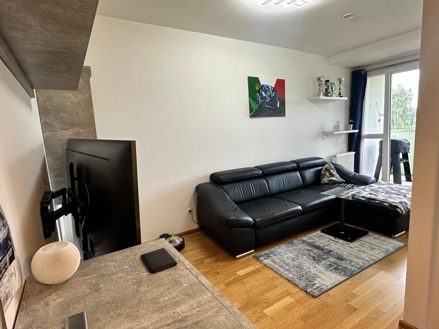 Wohnzimmer mit schwarzem Ledersofa, Holzboden und Zugang zu einem Balkon.