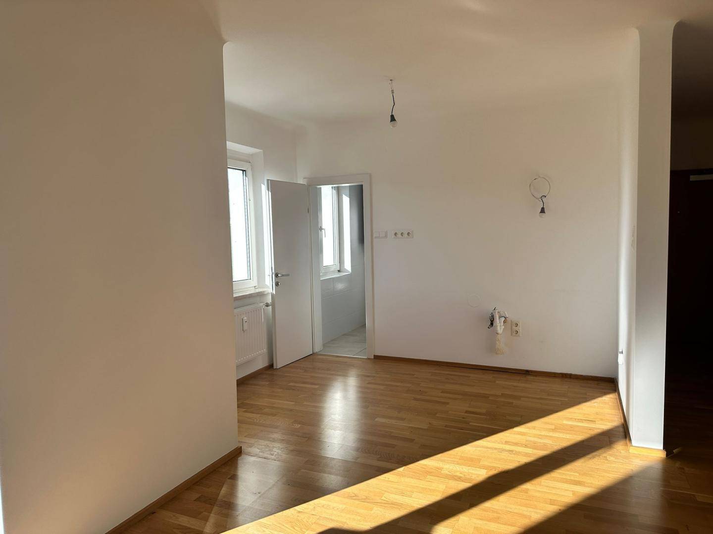Heller Wohnbereich mit Parkettboden und Zugang zu weiteren Räumen, durchflutet von Sonnenlicht.