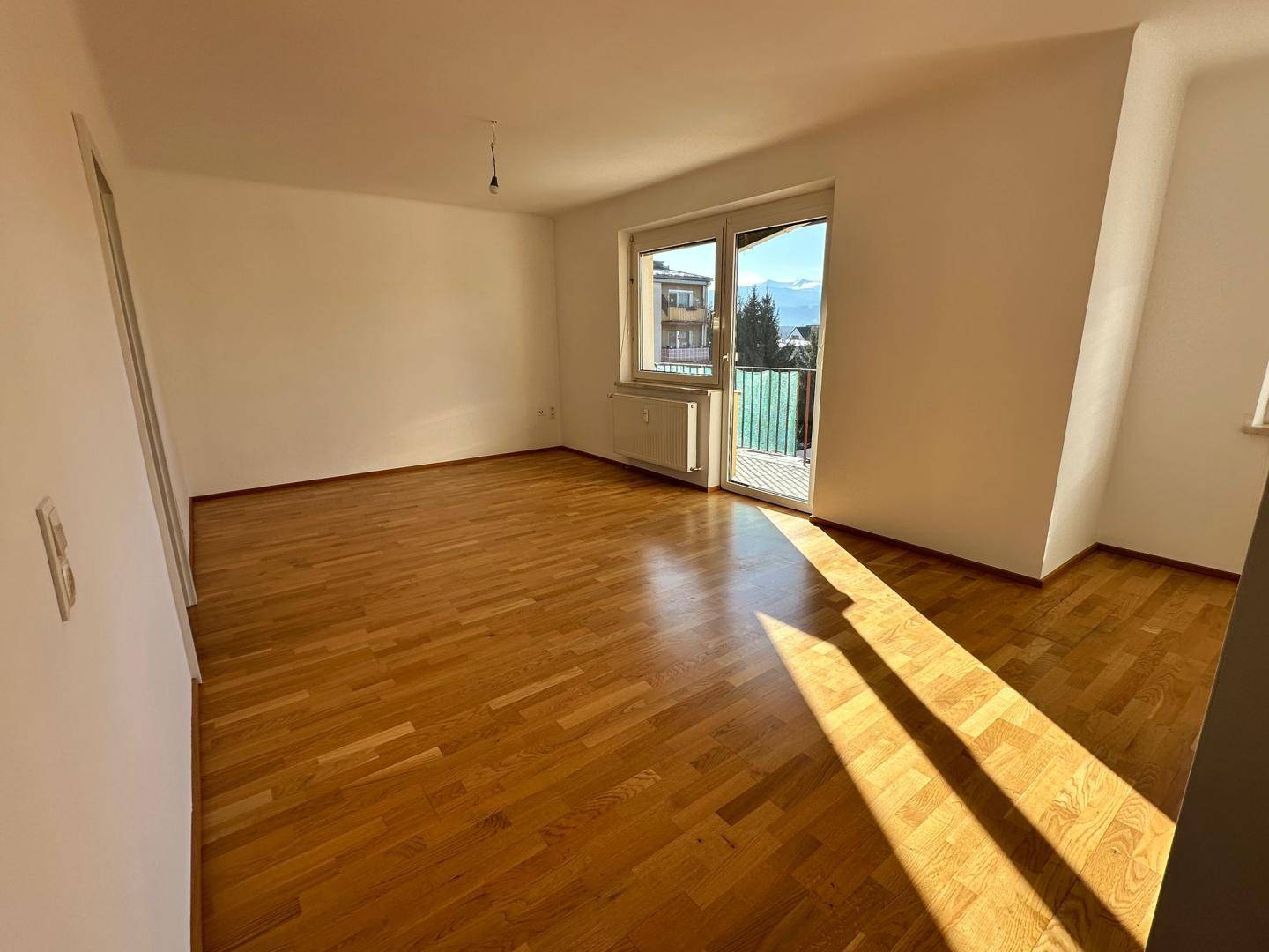 Geräumiges Wohnzimmer mit Parkettboden und Balkonzugang, durchflutet von hellem Sonnenlicht.