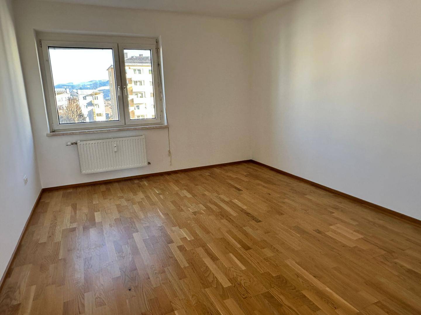 Leeres Schlafzimmer mit Parkettboden und einem Fenster, das einen Blick nach draußen ermöglicht.
