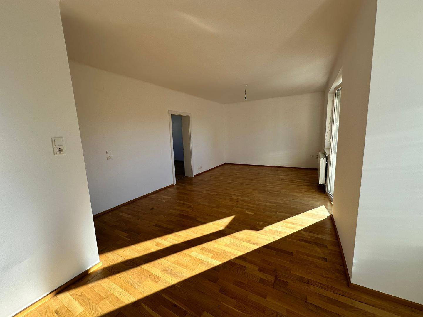 Geräumiges, leeres Wohnzimmer mit hellem Parkettboden und viel Tageslicht durch die Fenster.