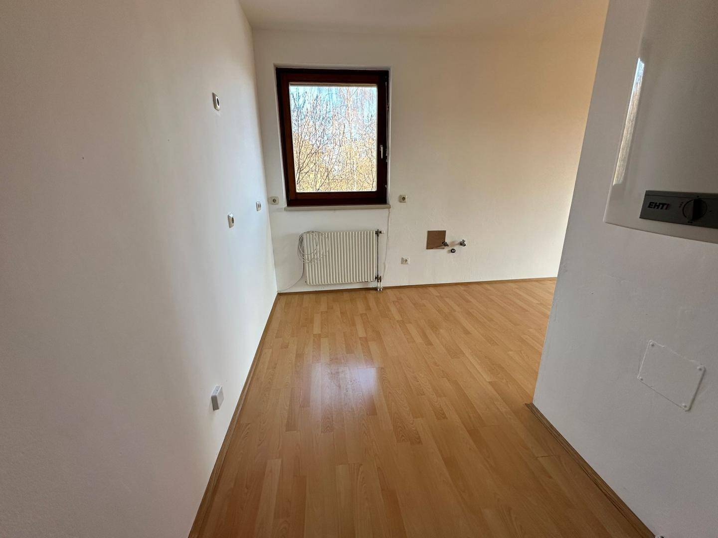 Helles, leeres Zimmer mit Fenster, Heizkörper und Anschlüssen an der Wand, ideal für verschiedene Nutzungen.