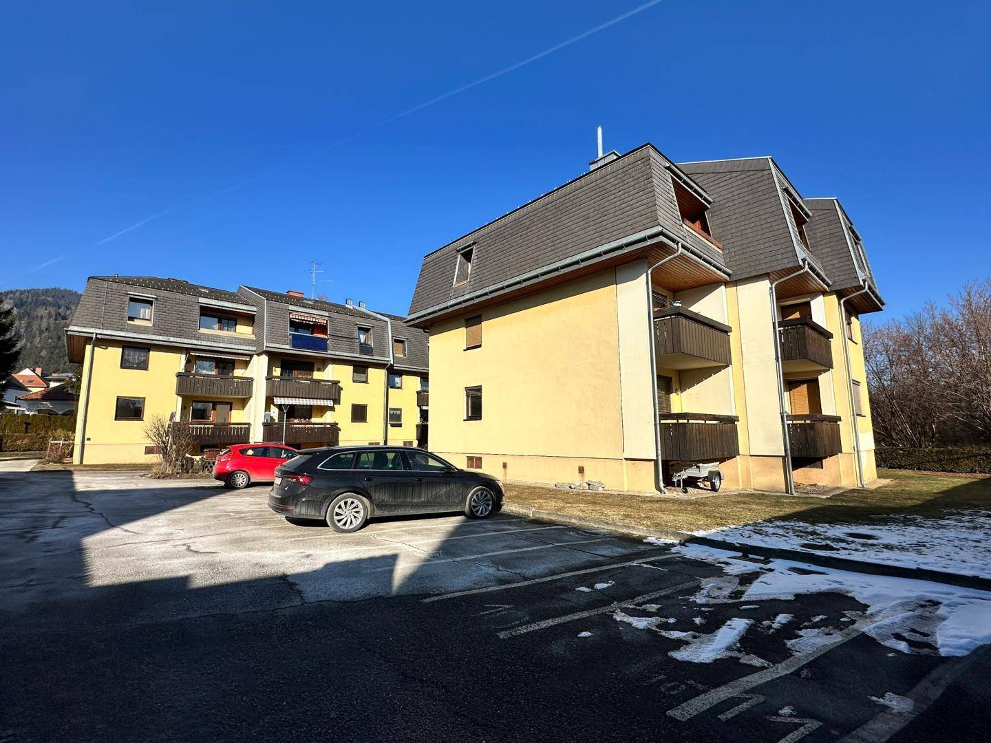 Mehrfamilienhäuser mit gelben Fassaden und Mansarddächern, umgeben von einem Parkplatz und Grünflächen.