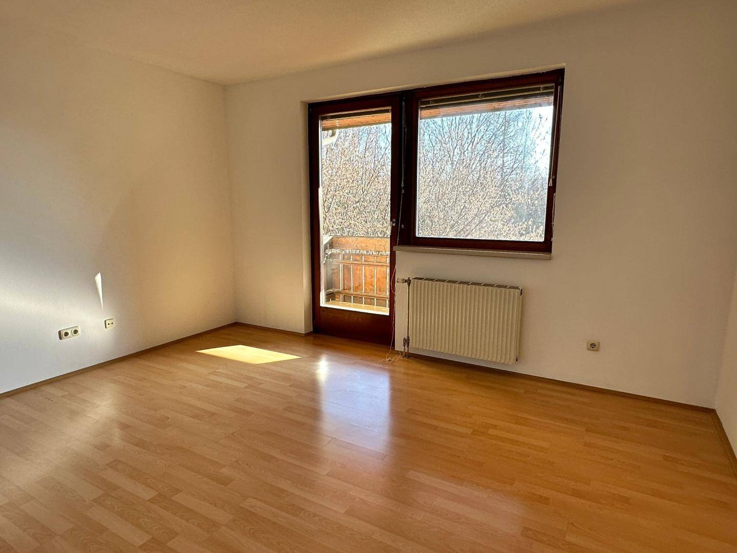 Helles Wohnzimmer mit Holzboden und großem Fenster, das Zugang zu einem Balkon bietet.