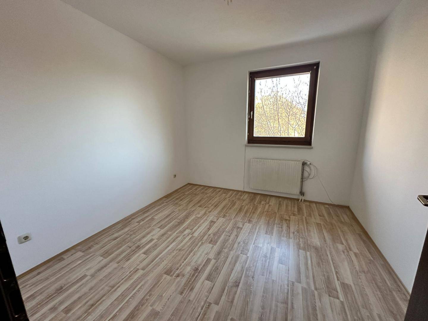 Gemütliches, leeres Schlafzimmer mit Laminatboden, weiß gestrichenen Wänden und einem Fenster mit Heizkörper.