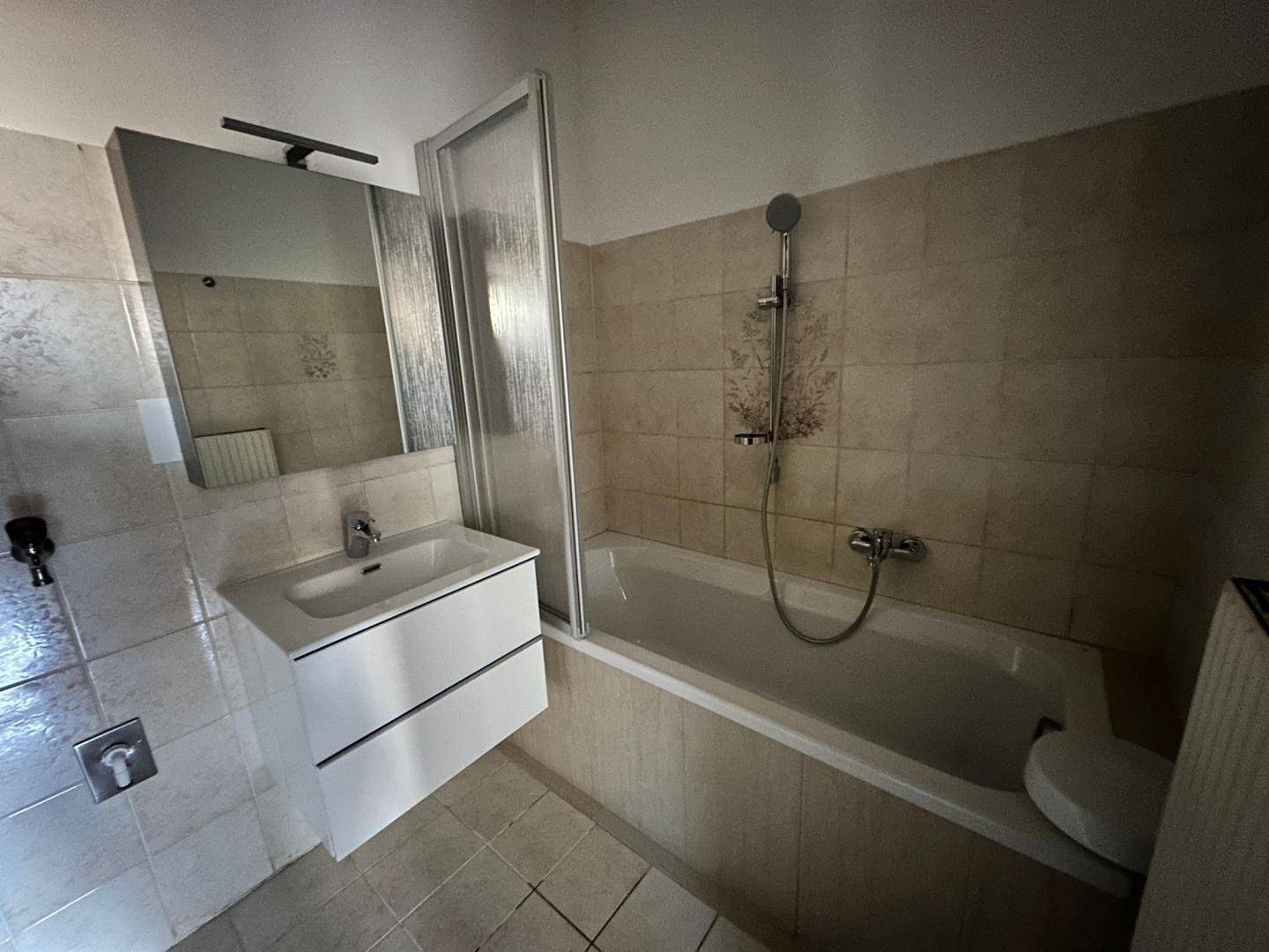Badezimmer mit beigefarbenen Fliesen, Badewanne mit Duschwand und modernem Waschtisch mit Spiegelschrank.