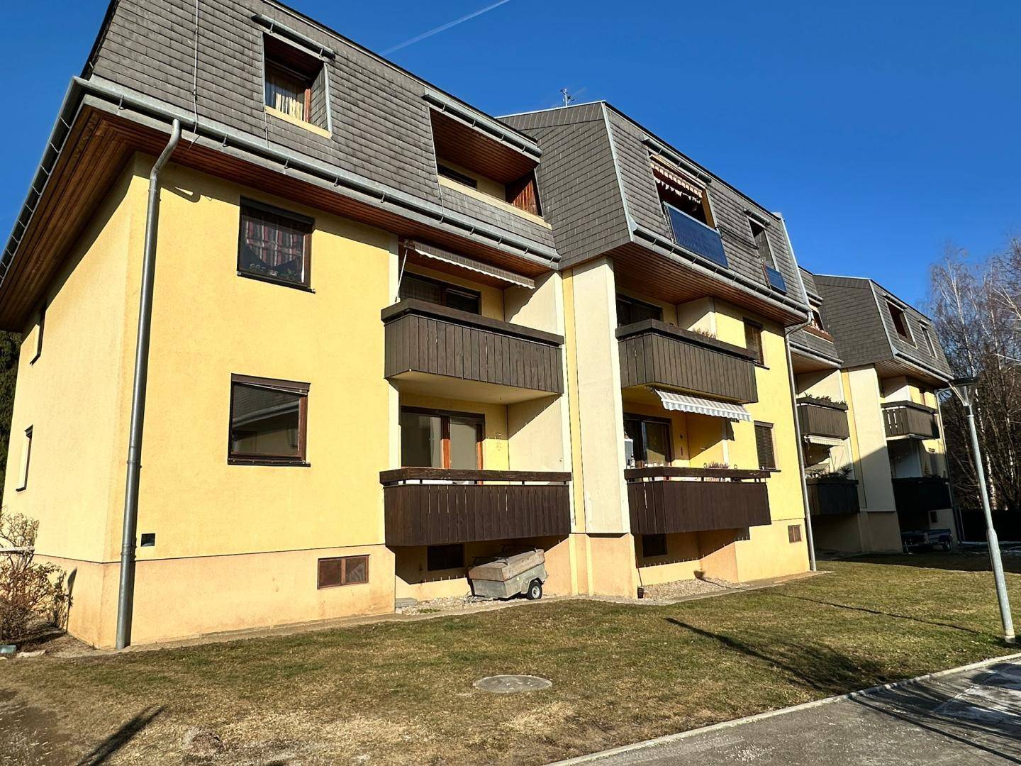 Mehrfamilienhaus mit gelber Fassade, braunen Balkonen und Mansarddach in sonniger Umgebung.