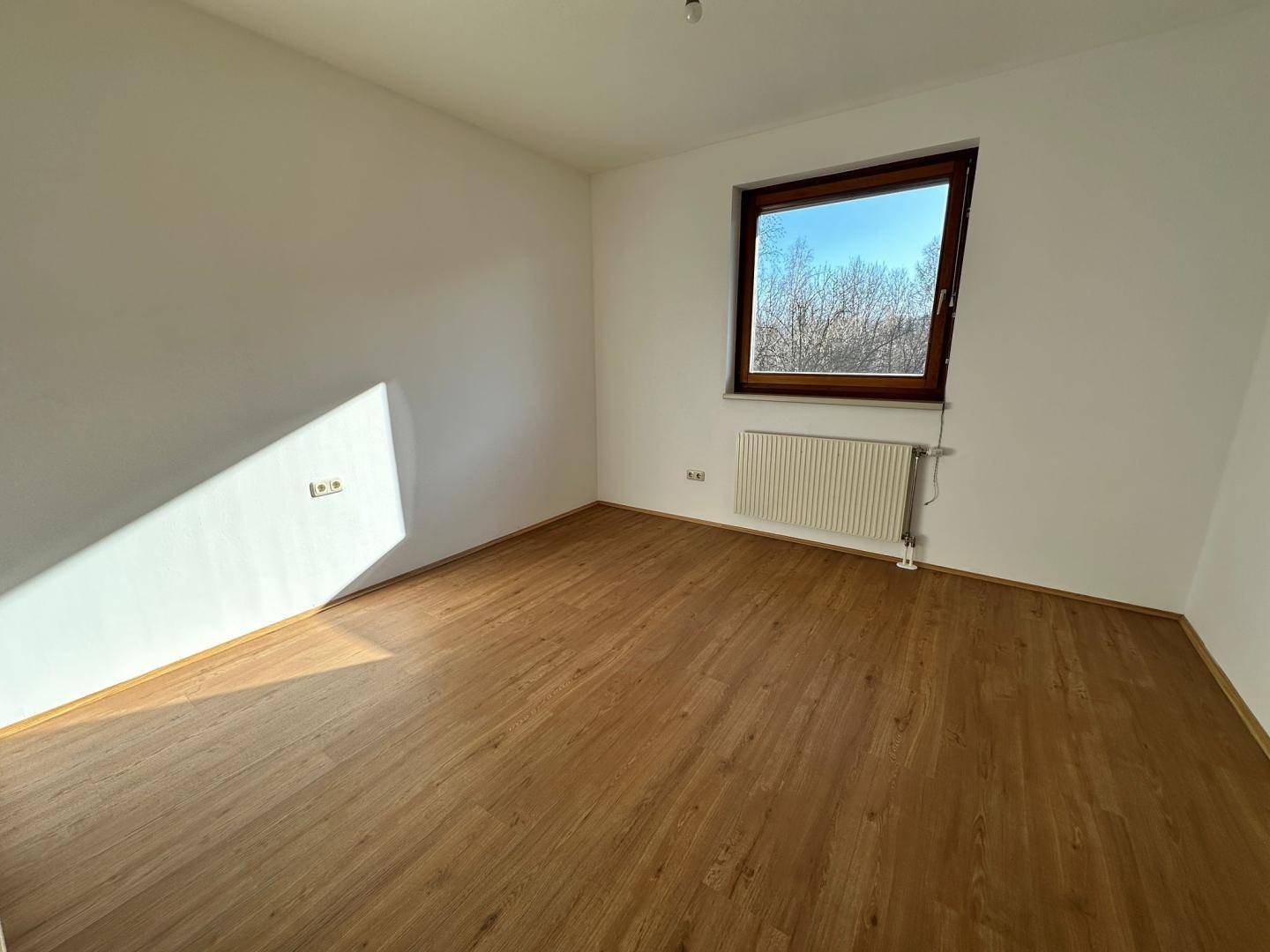 Helles Schlafzimmer mit Laminatboden und einem großen Fenster, das viel Tageslicht hereinlässt.