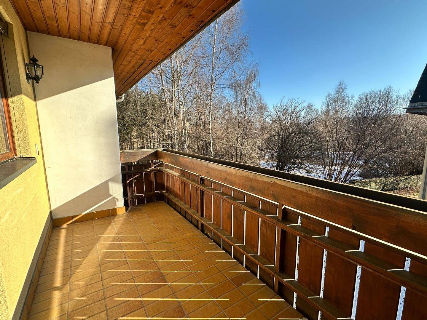 Geräumiger Balkon mit Fliesenboden und Holzgeländer, umgeben von Bäumen und Natur.