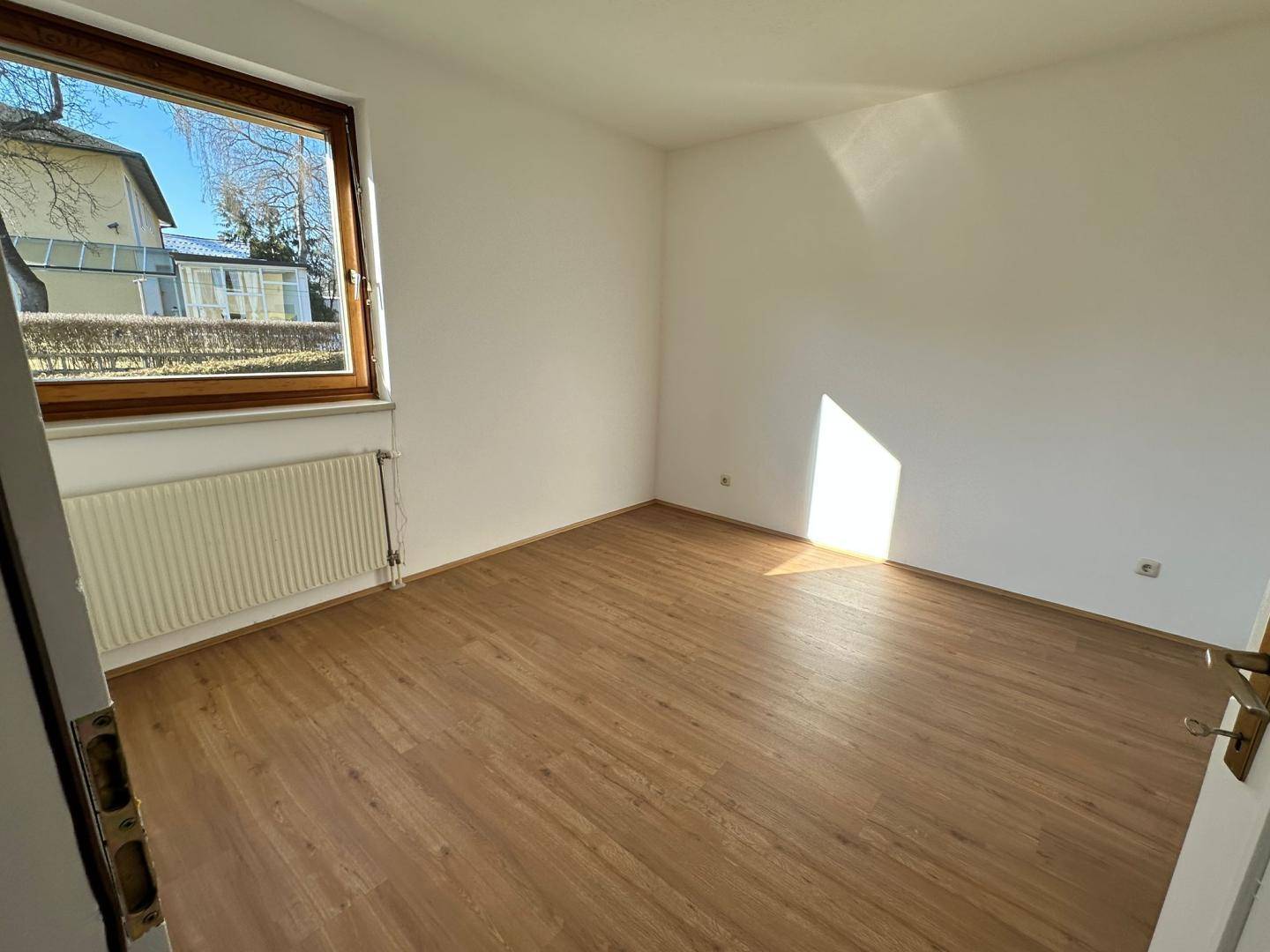 Helles Zimmer mit Laminatboden und großem Fenster, das viel Tageslicht hereinlässt.