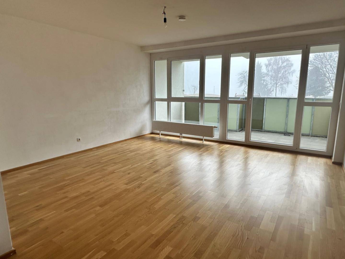 Heller Wohnraum mit Parkettboden und breiter Fensterfront, die viel Tageslicht hereinlässt.