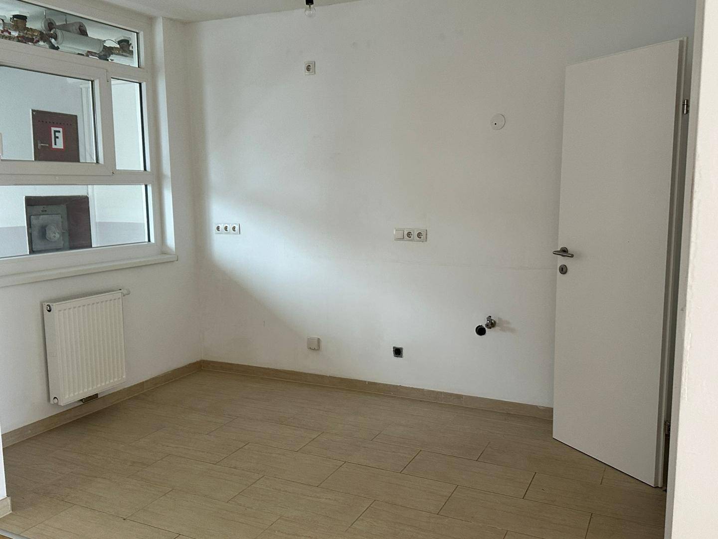 Leerer Küchenraum mit Fliesenboden, Fenster und Anschlüssen für eine Einbauküche.