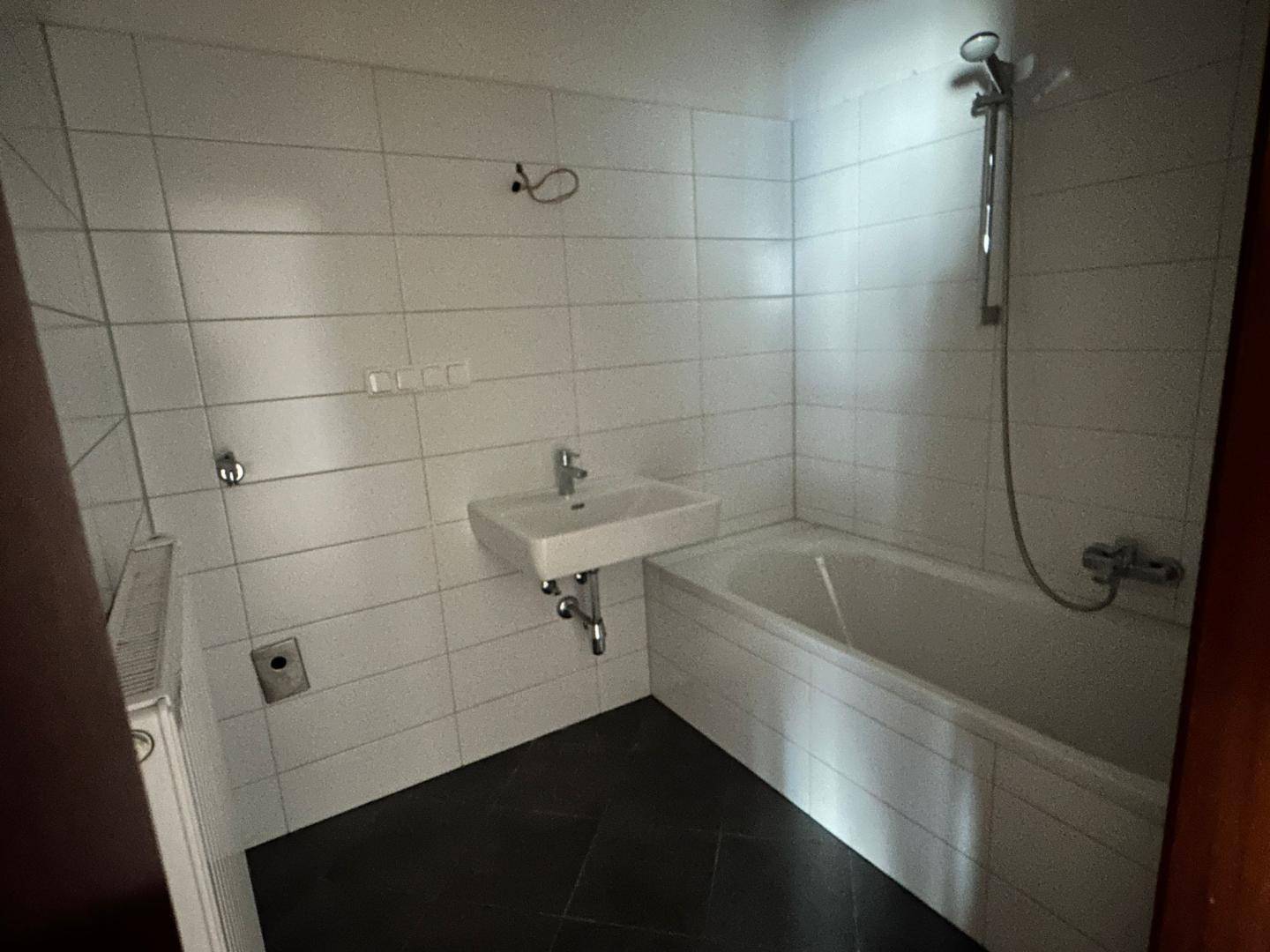 Badezimmer mit weißen Wandfliesen, dunklem Boden, Badewanne mit Duschfunktion und Waschbecken.