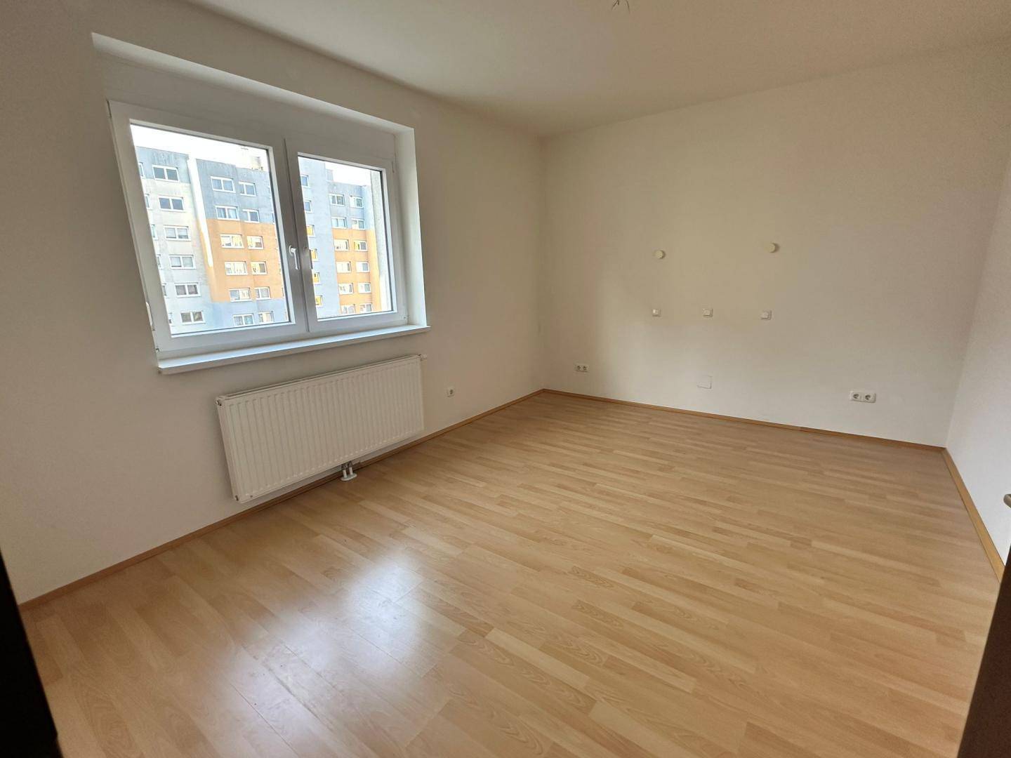 Geräumiges Schlafzimmer mit hellem Laminatboden und großem Fenster mit Blick auf die umliegenden Gebäude.