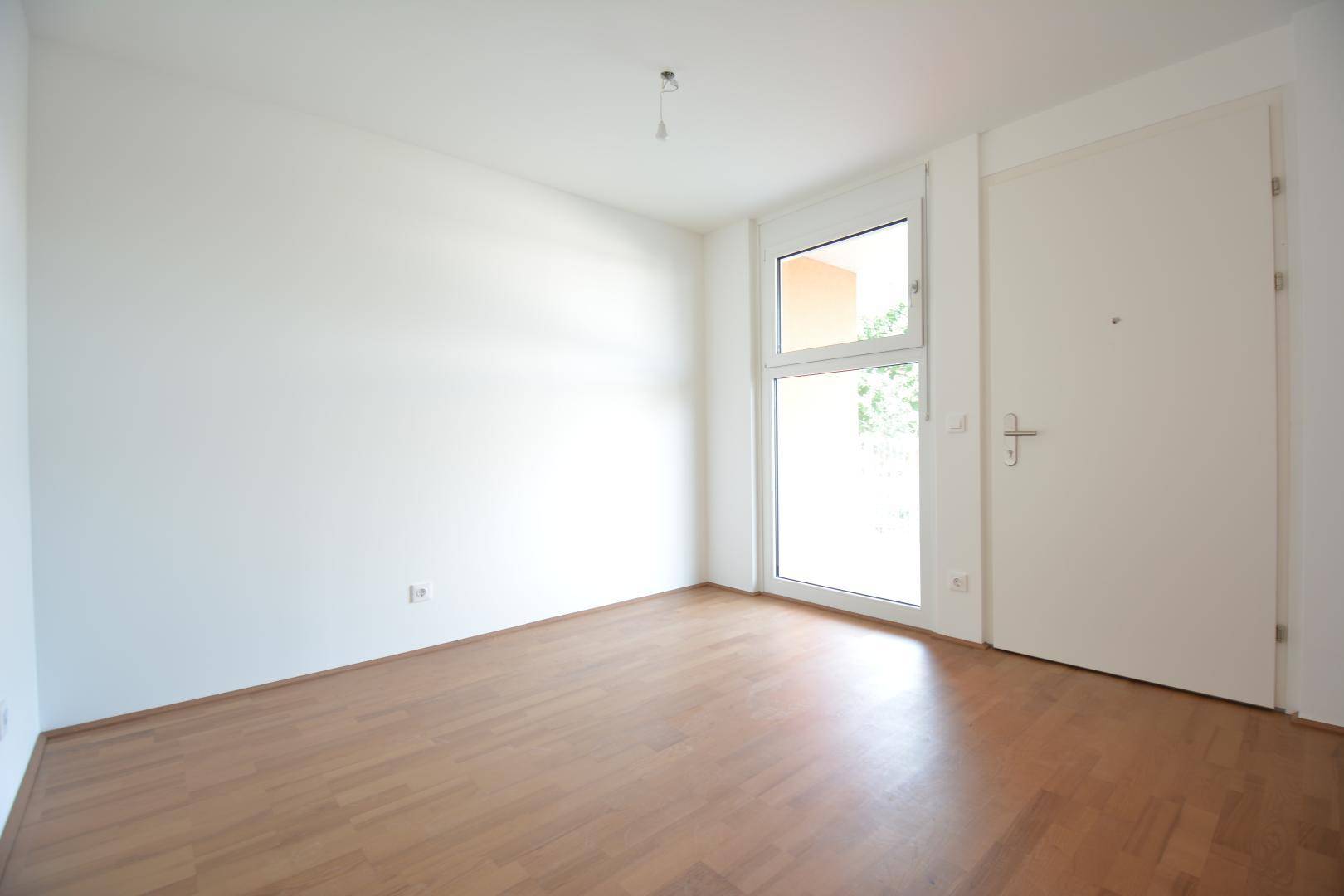 Leerer Raum mit hellem Holzfußboden und einer großen Balkontür für viel Tageslicht.