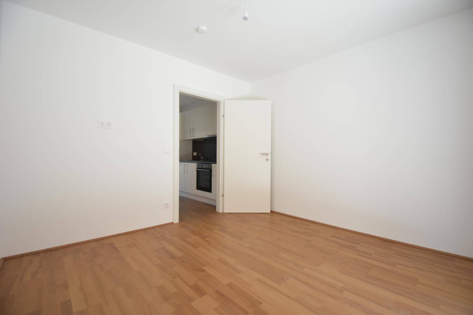 Leerer Wohnbereich mit Holzboden und offener Tür zur angrenzenden, modernen Küche.