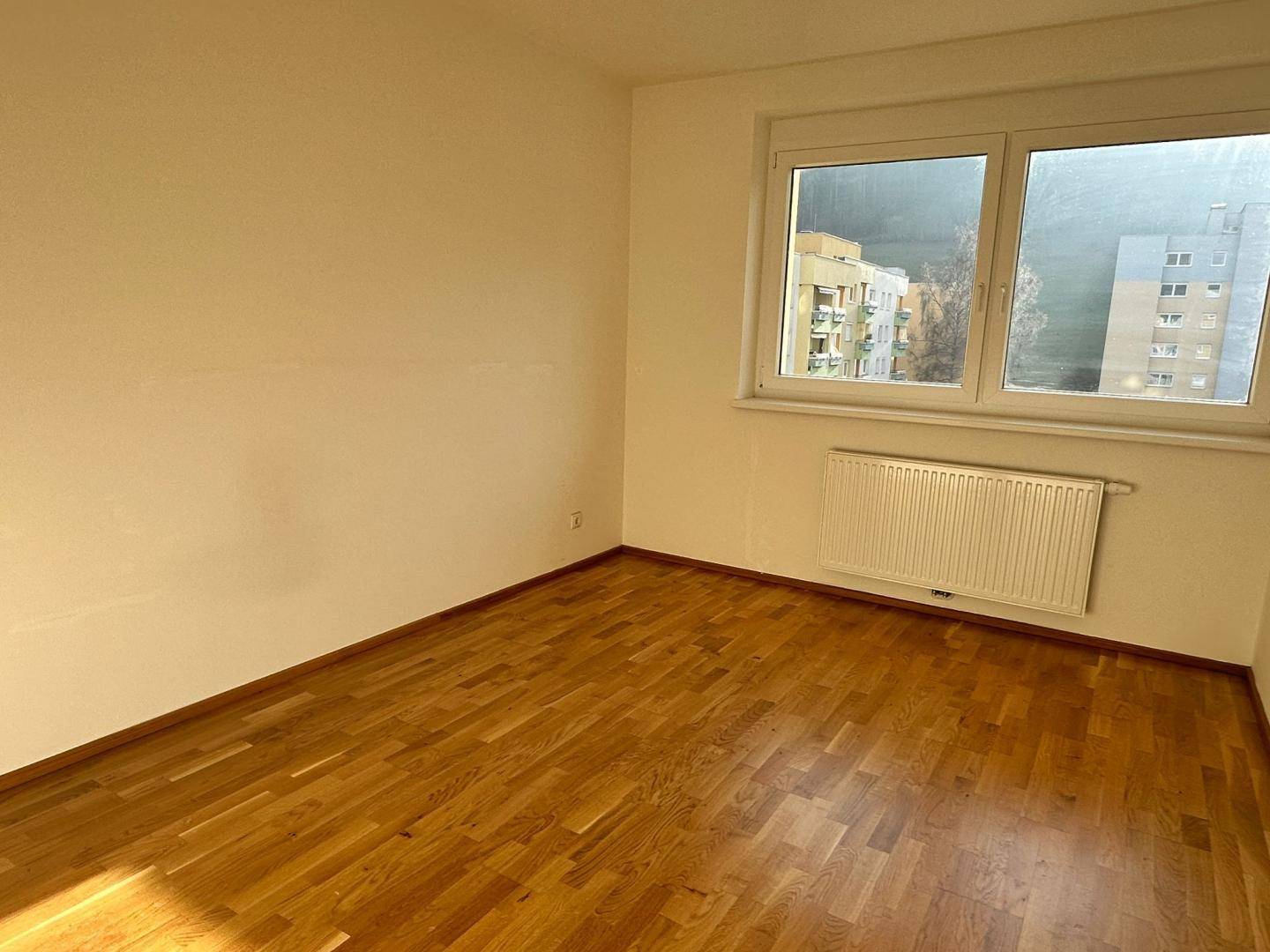 Leeres Zimmer mit Holzboden, Fenster und Blick auf umliegende Gebäude und Berge.