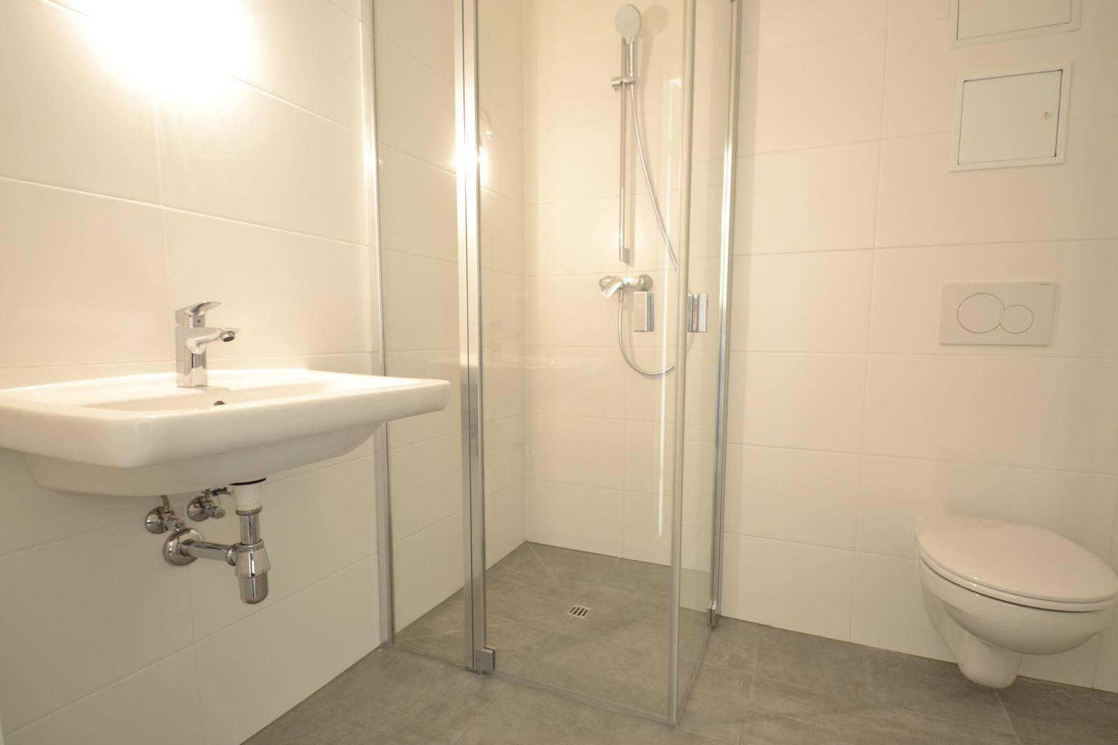 Modernes Badezimmer mit weißen Fliesen, Waschbecken, wandhängendem WC und begehbarer Dusche.