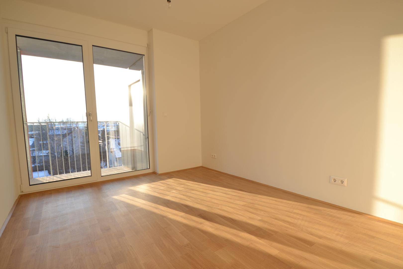 Helles Schlafzimmer mit Parkettboden und großem Fenster, das viel Tageslicht hereinlässt.