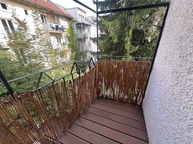 Kleiner Balkon mit Holzboden und Sichtschutz, umgeben von Bäumen und Blick auf Nachbargebäude.