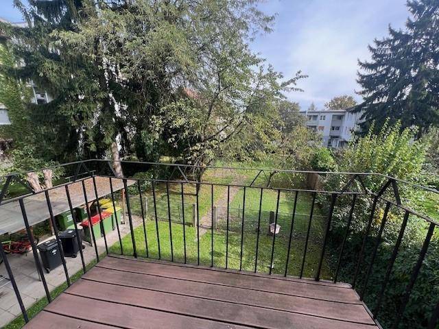 Balkon mit Holzboden und Blick auf einen grünen Garten mit Bäumen und umliegenden Gebäuden.