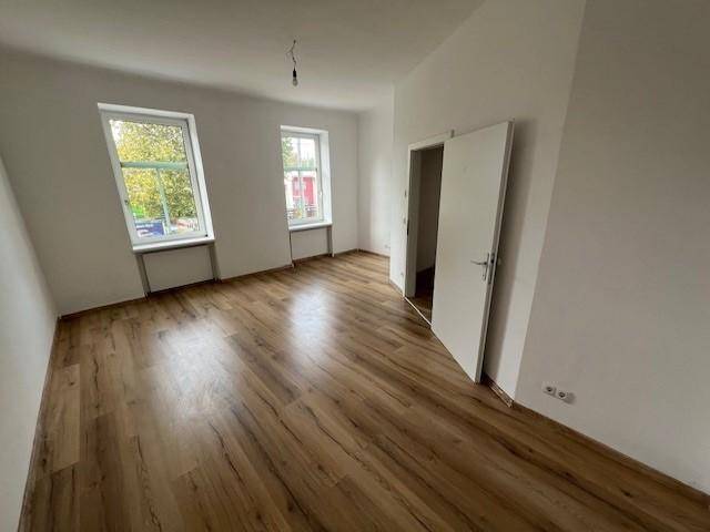 Helles Wohnzimmer mit zwei Fenstern, Laminatboden und weiß gestrichenen Wänden.