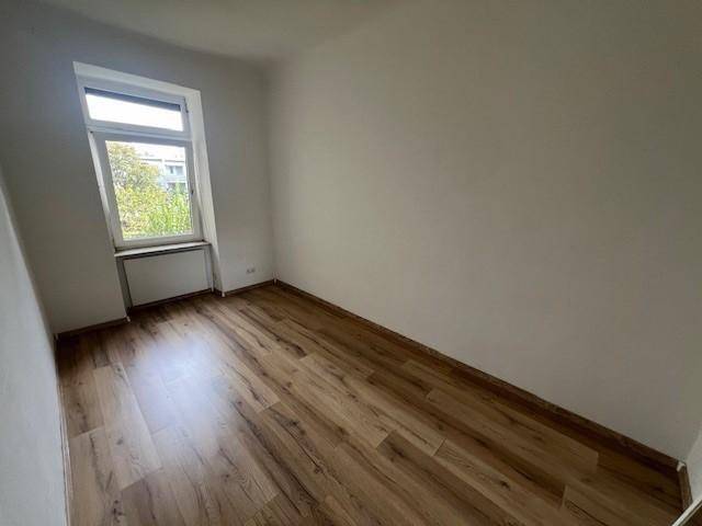 Helles, leeres Zimmer mit Laminatboden und einem Fenster, das viel Tageslicht hereinlässt.
