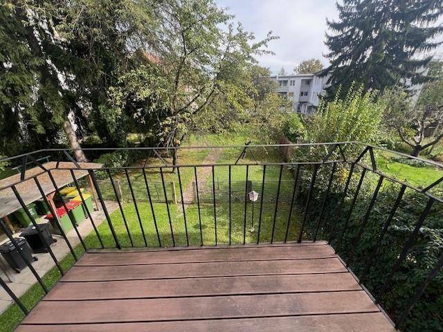 Balkon mit Holzboden und weitem Blick über einen grünen Gartenbereich mit altem Baumbestand.