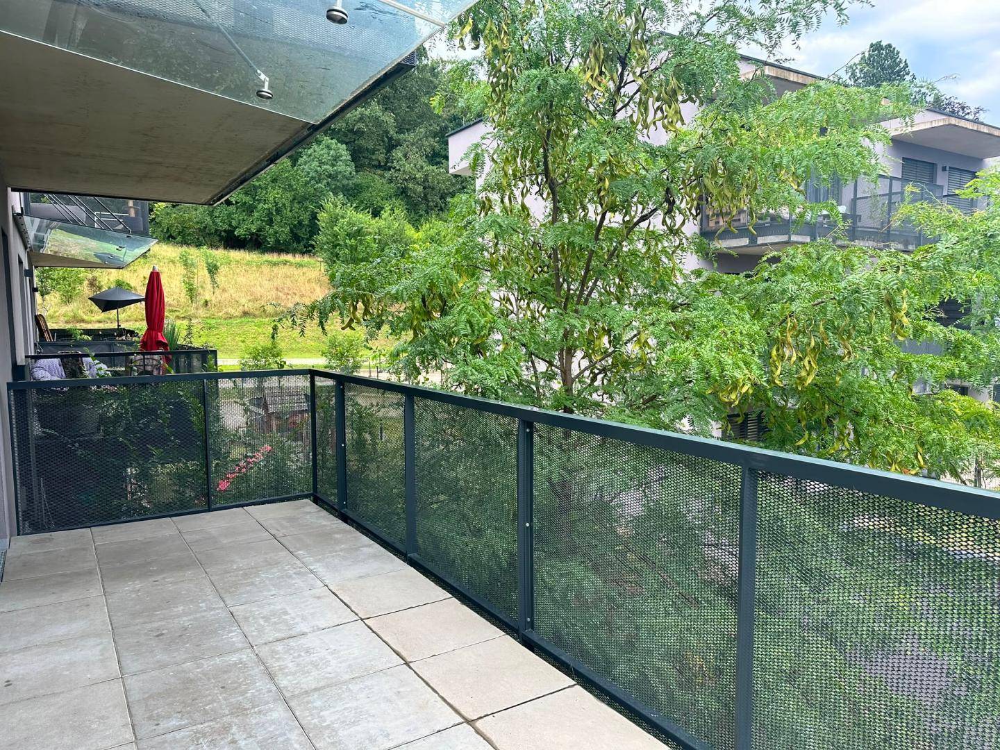 Geräumiger Balkon mit Fliesenboden und Metallgeländer, umgeben von viel Grün.