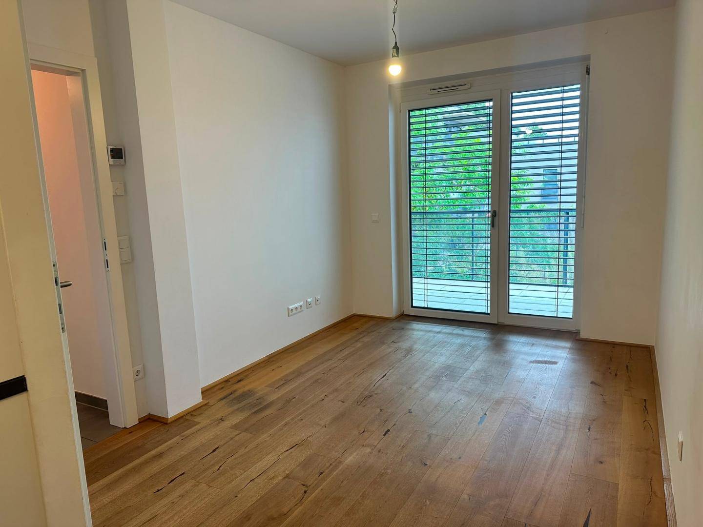 Helles Zimmer mit Holzboden und großen Fenstern mit Jalousien, bietet Zugang zum Außenbereich.
