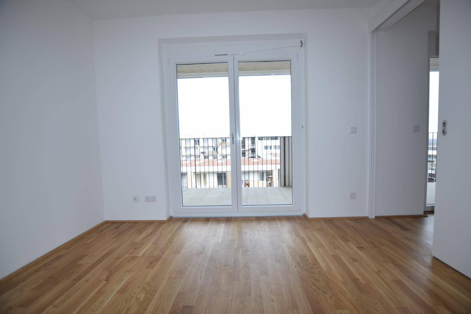 Geräumiger Raum mit hellem Holzboden und breiter Balkontür, die einen Blick nach draußen ermöglicht.