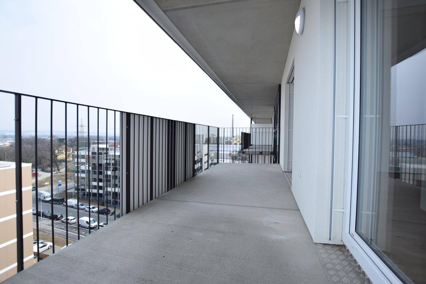 Langer Balkon mit Betonboden und schwarzem Geländer, bietet einen weiten Blick über die Stadt.