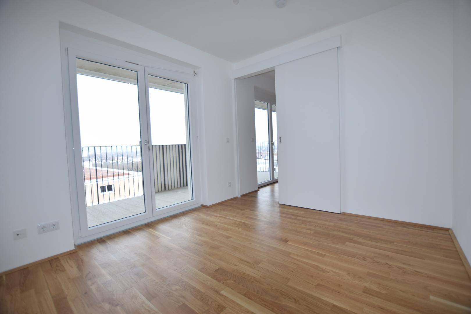 Heller Wohnraum mit Holzboden, Balkonzugang und einer modernen Schiebetür zu einem weiteren Zimmer.