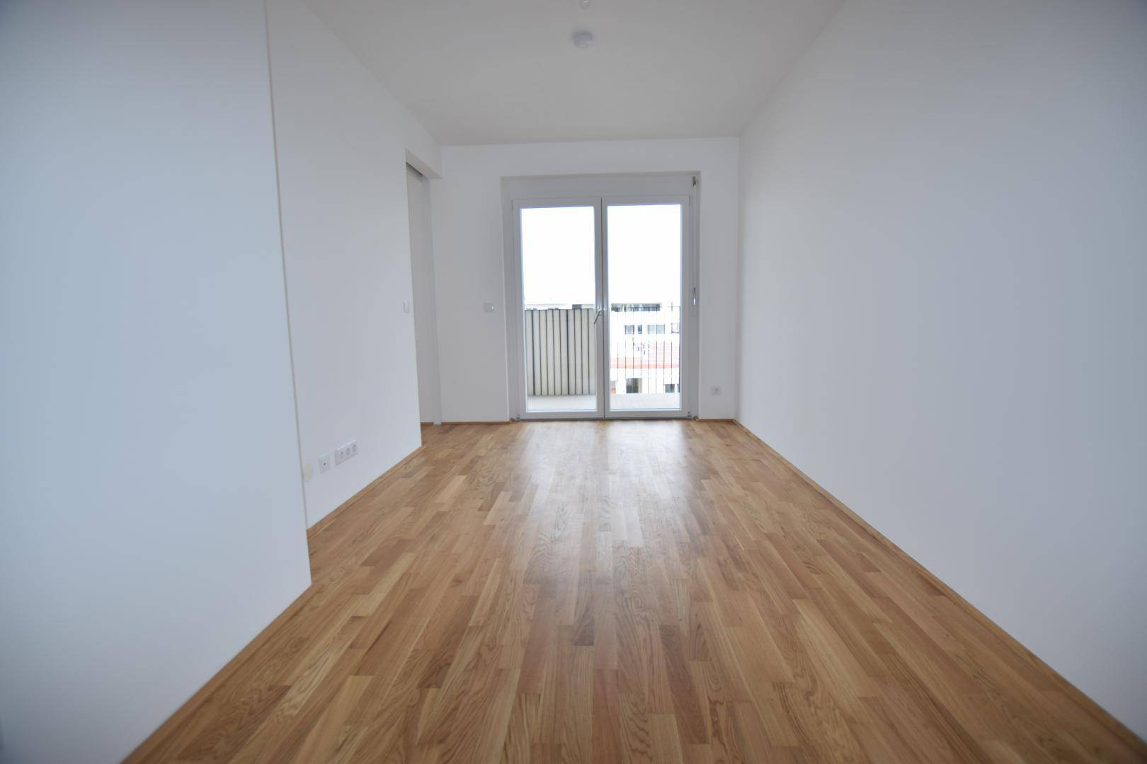 Heller Raum mit Holzboden und Zugang zu einem kleinen Balkon, der viel Tageslicht hereinlässt.