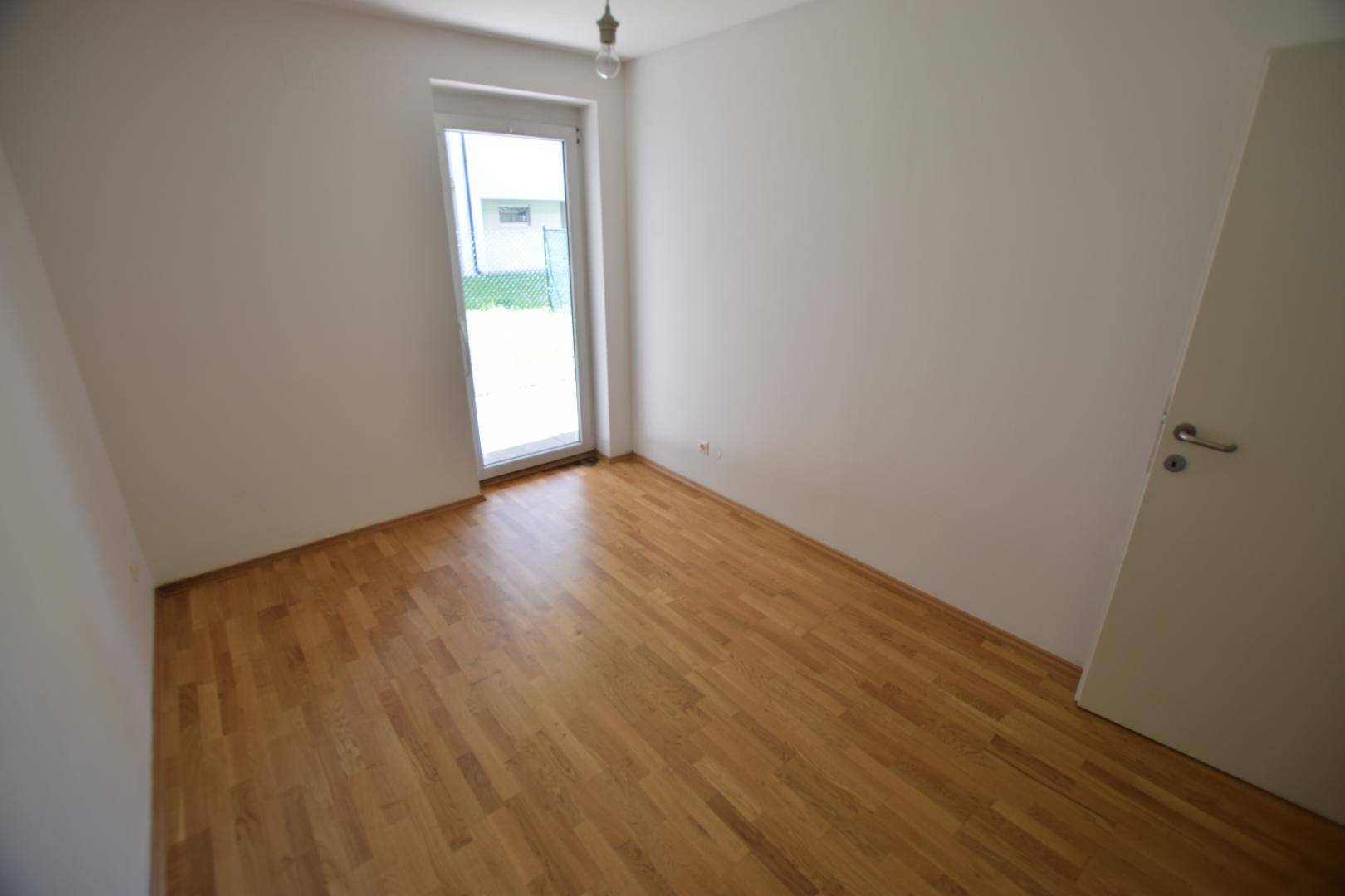 Freundliches Zimmer mit Holzboden und großer Glastür, die viel Tageslicht hereinlässt.