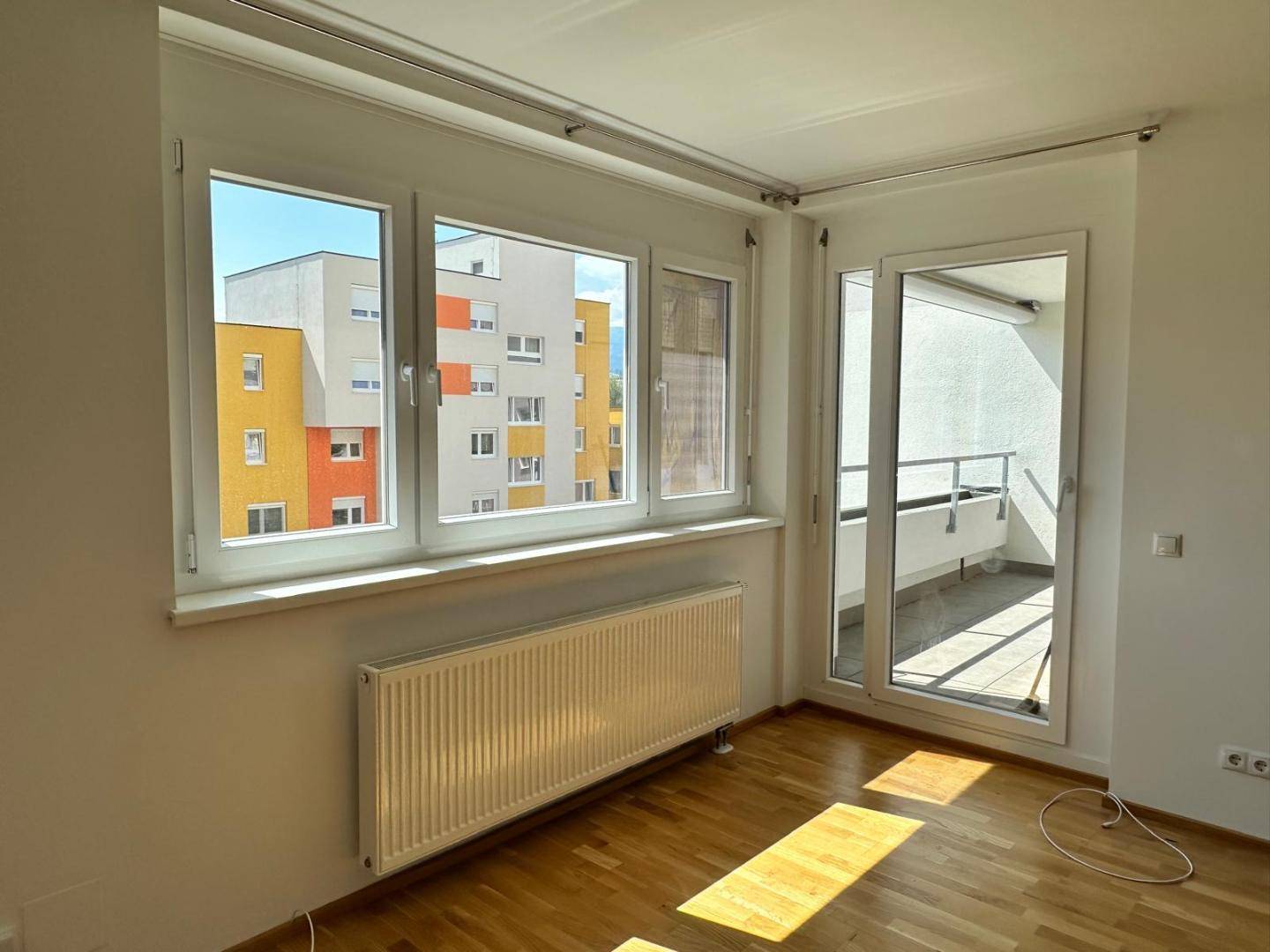 Helles Wohnzimmer mit Parkettboden, großem Fenster und Zugang zu einem sonnigen Balkon.