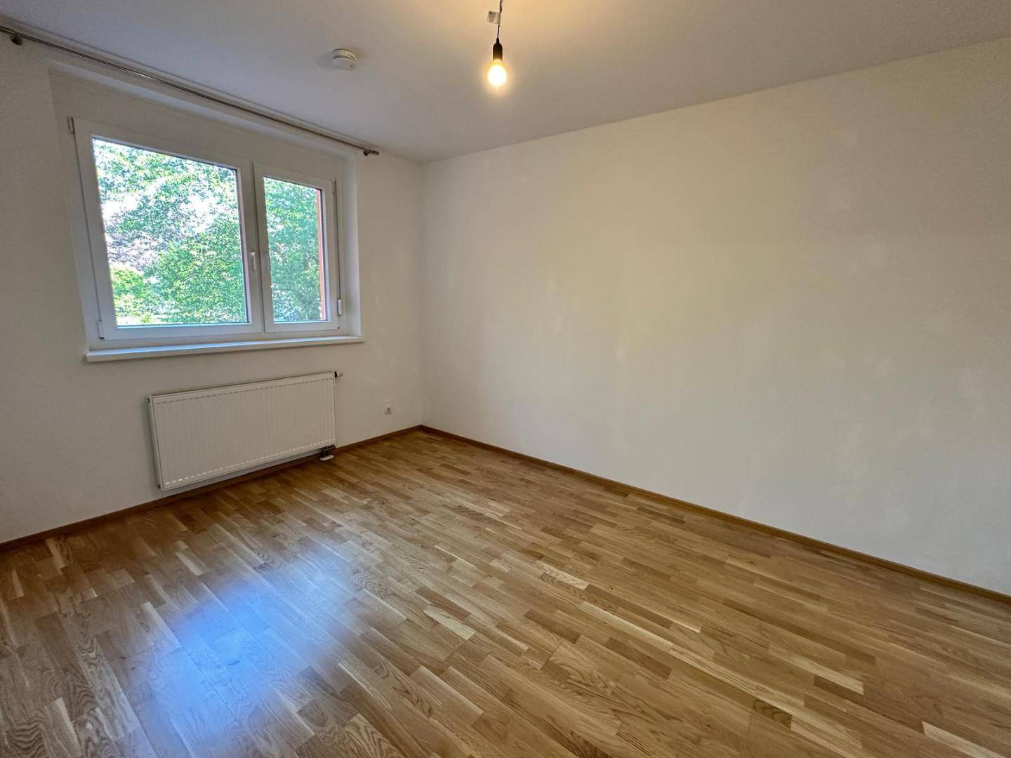 Helles Schlafzimmer mit Parkettboden und Fenster, das einen Blick ins Grüne bietet.