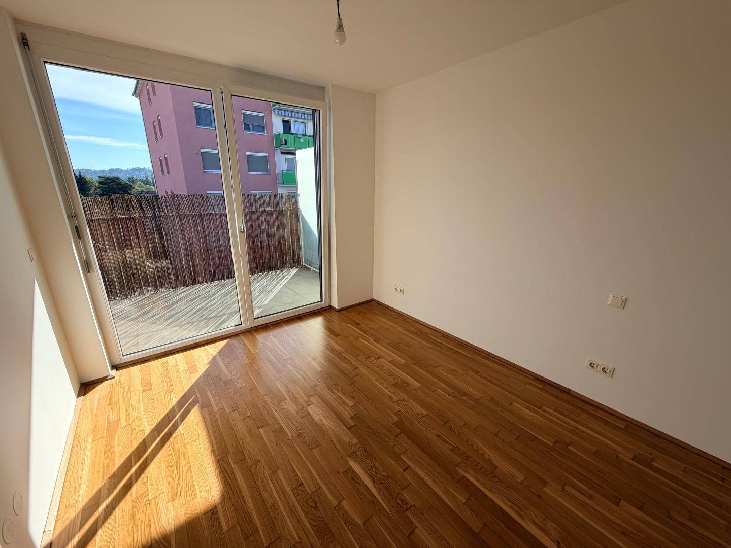Lichtdurchfluteter Wohnraum mit Holzboden und großem Fenster mit Zugang zum Balkon.