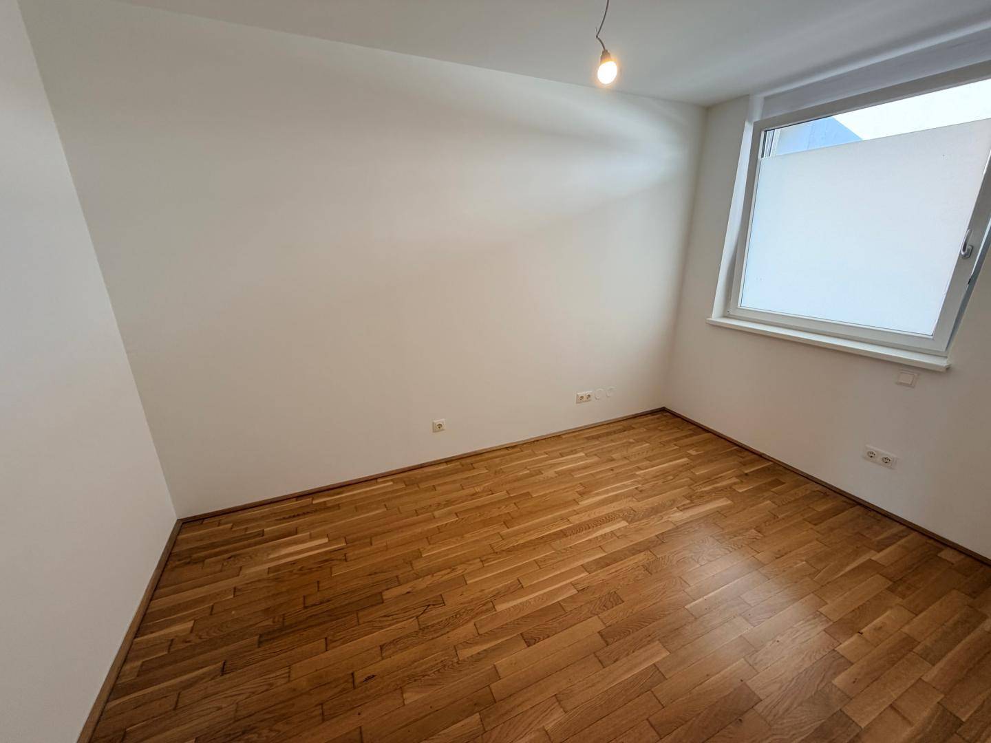 Helles Schlafzimmer mit Parkettboden und einem Fenster, das viel Tageslicht hereinlässt.