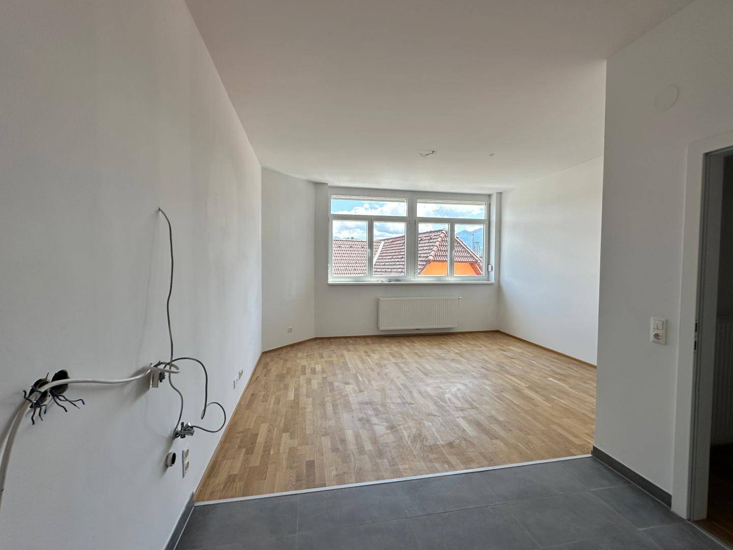Lichtdurchfluteter Wohnraum mit großen Fenstern, Parkettboden und Blick auf Dächer.
