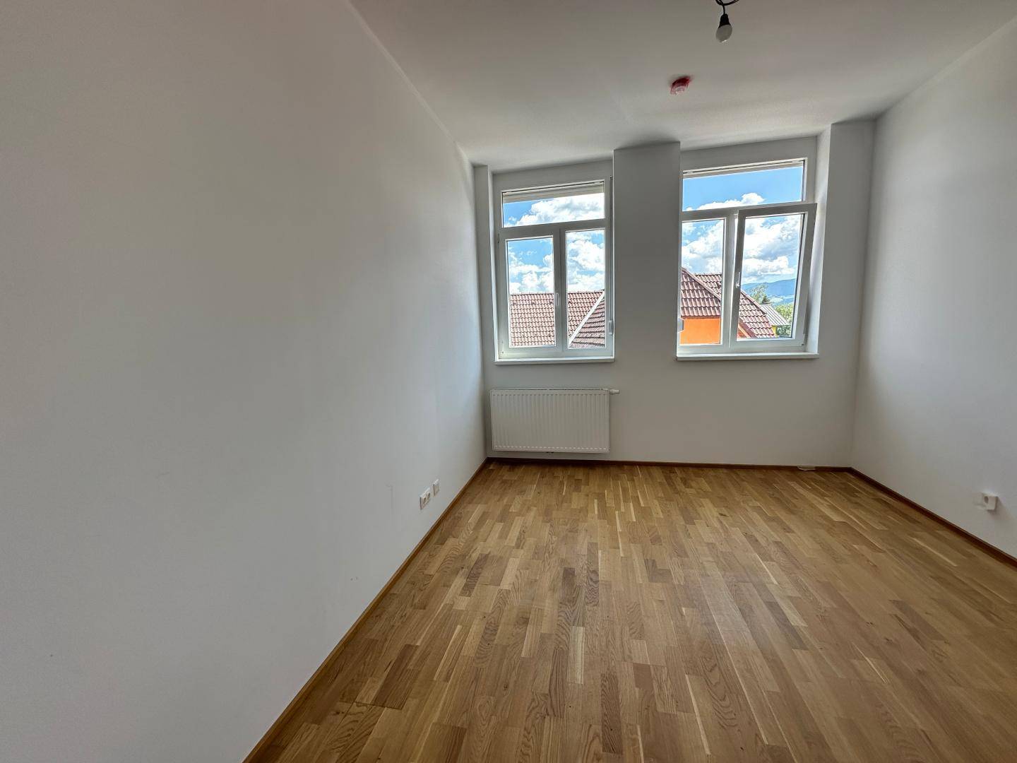 Helles Schlafzimmer mit zwei Fenstern, Parkettboden und Blick auf die umliegenden Dächer.
