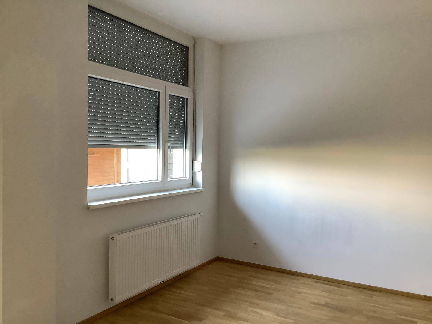 Leeres Zimmer mit hellem Parkettboden, weiß gestrichenen Wänden und einem Fenster mit Rollladen.