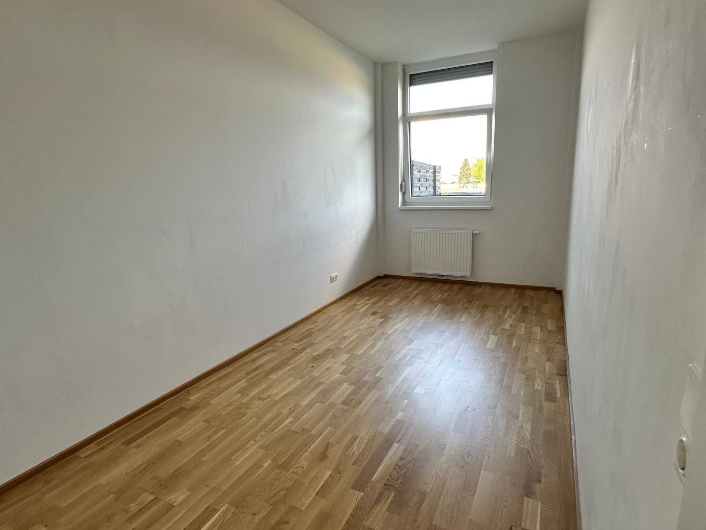 Helles, leeres Zimmer mit Parkettboden, weißen Wänden und einem Fenster mit Blick nach draußen.