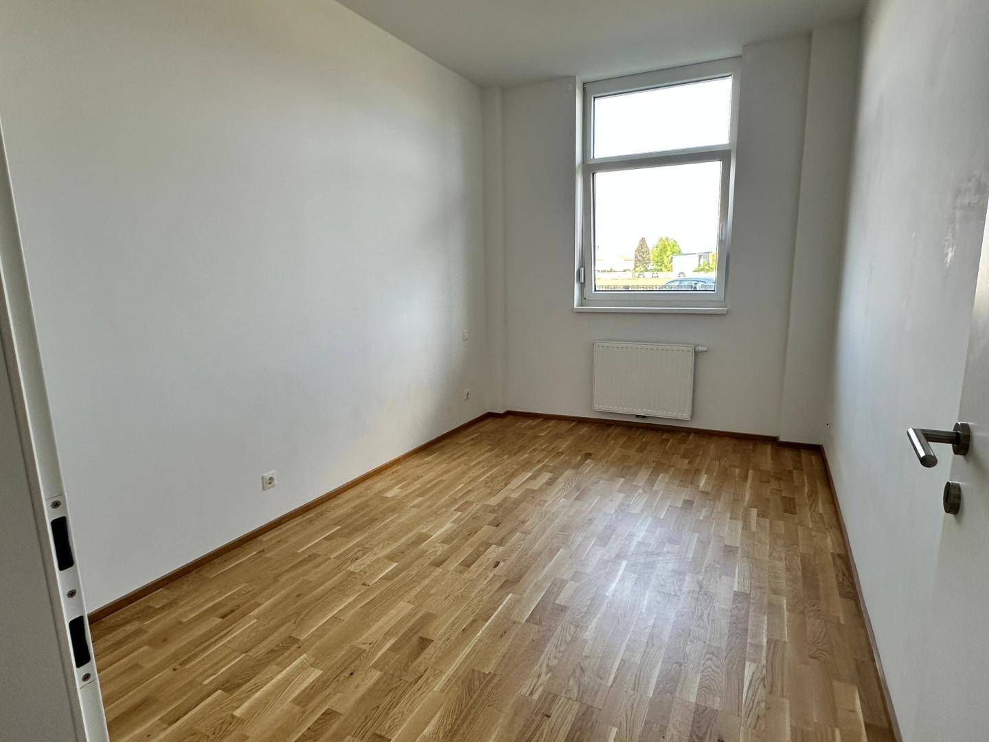 Leeres Zimmer mit hellem Parkettboden, weiß gestrichenen Wänden und einem Fenster.