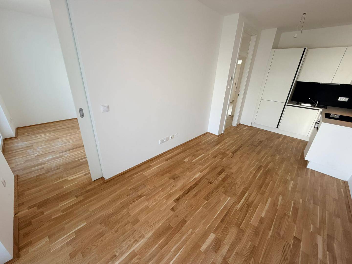 Offener Wohnbereich mit Holzboden, der zur Küche und einem weiteren Zimmer führt.