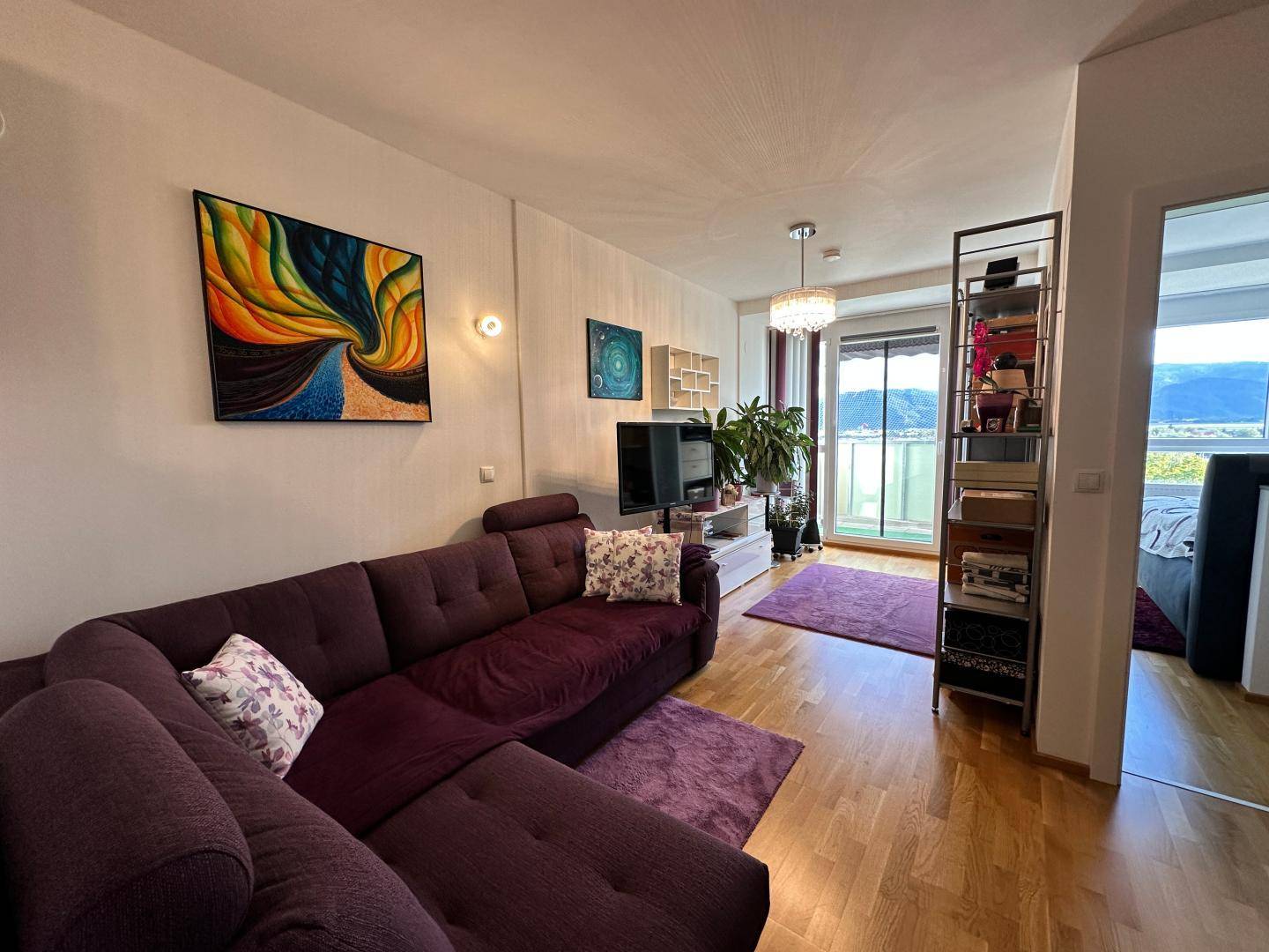 Gemütliches Wohnzimmer mit lila Ecksofa, Holzboden und Zugang zu einem Balkon mit Bergblick.