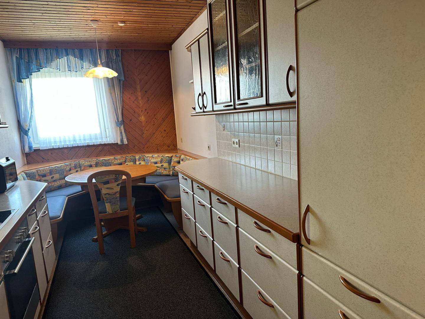 Gemütliche Küche mit Holzvertäfelung, Essbereich und einem Fenster mit traditionellen Vorhängen.