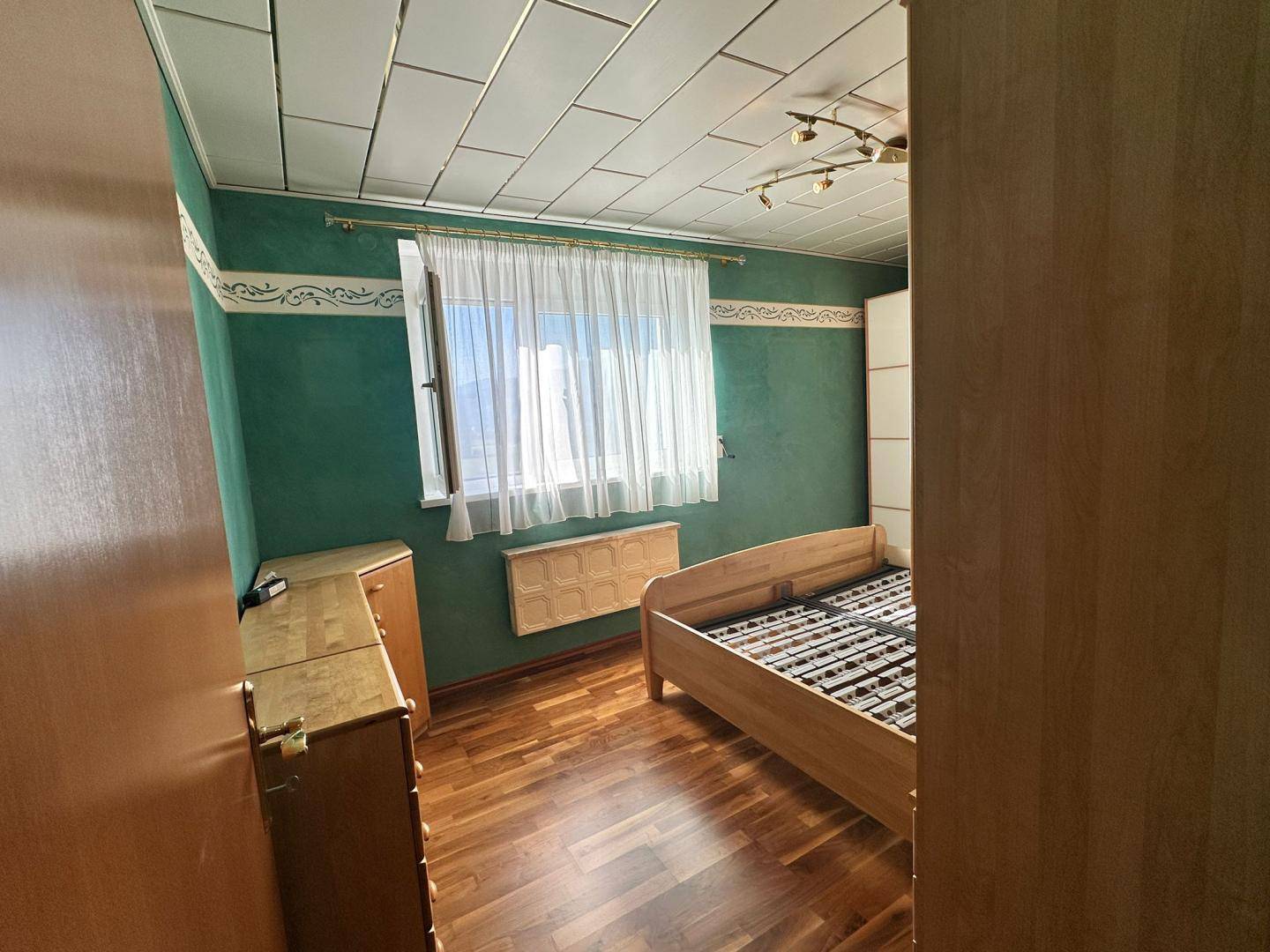 Schlafzimmer mit grünen Wänden, Holzboden und einem Fenster, das viel natürliches Licht hereinlässt.