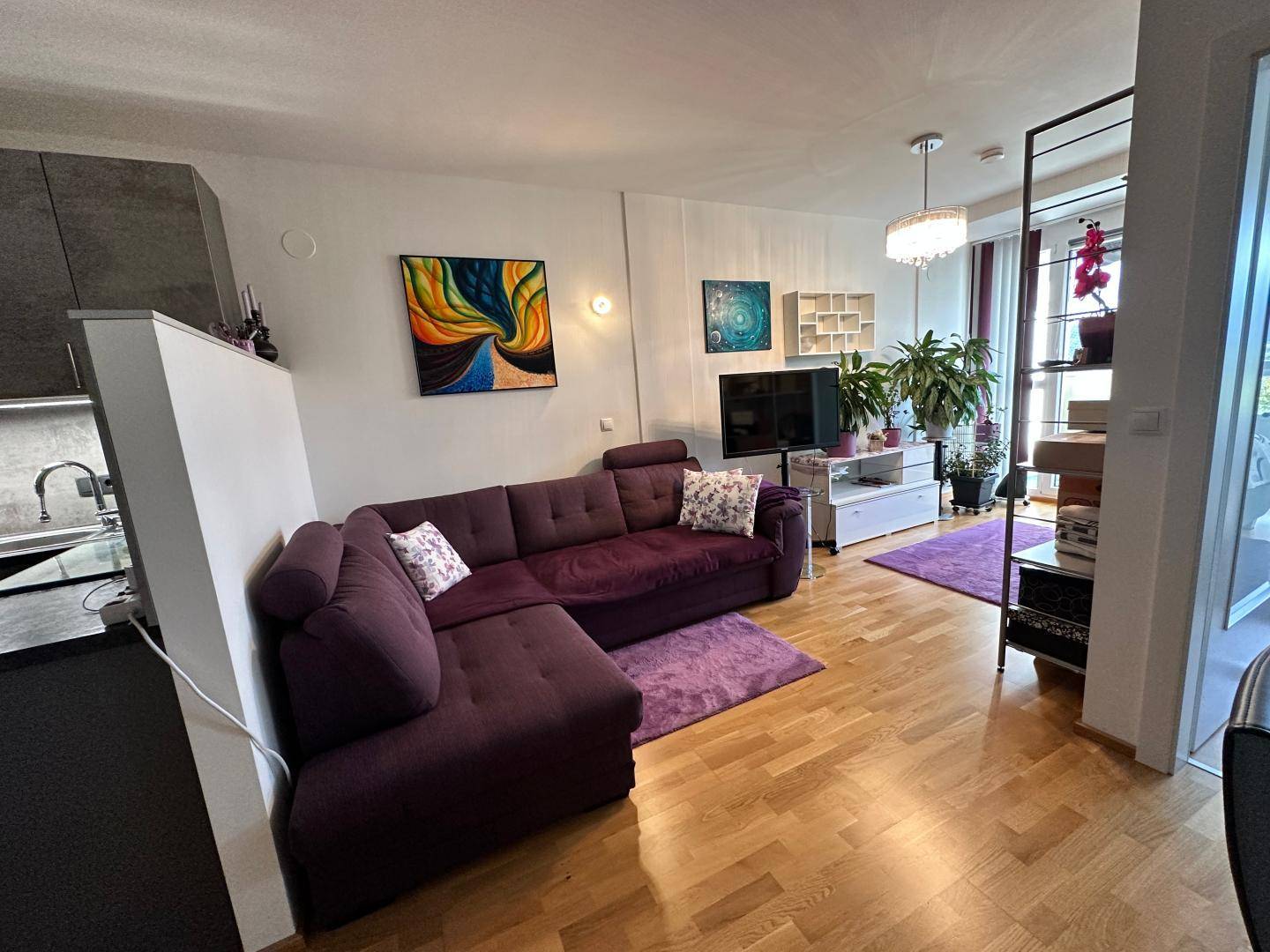 Geräumiges Wohnzimmer mit lila Ecksofa, farbenfrohen Bildern und Holzboden.